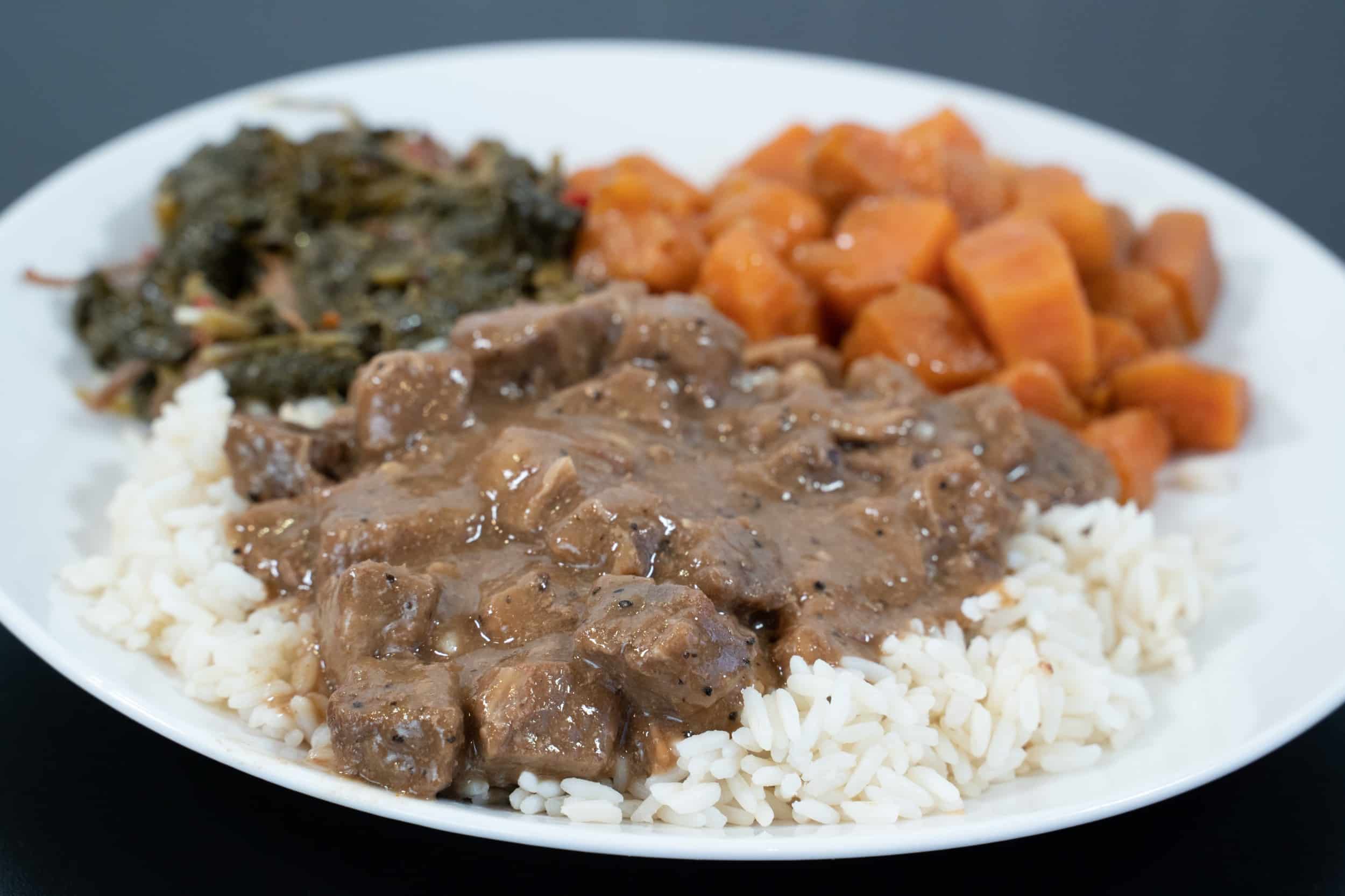 Plate of white rice topped with beef in brown gravy, plus roasted sweet potatoes and sautéed greens on a white plate.