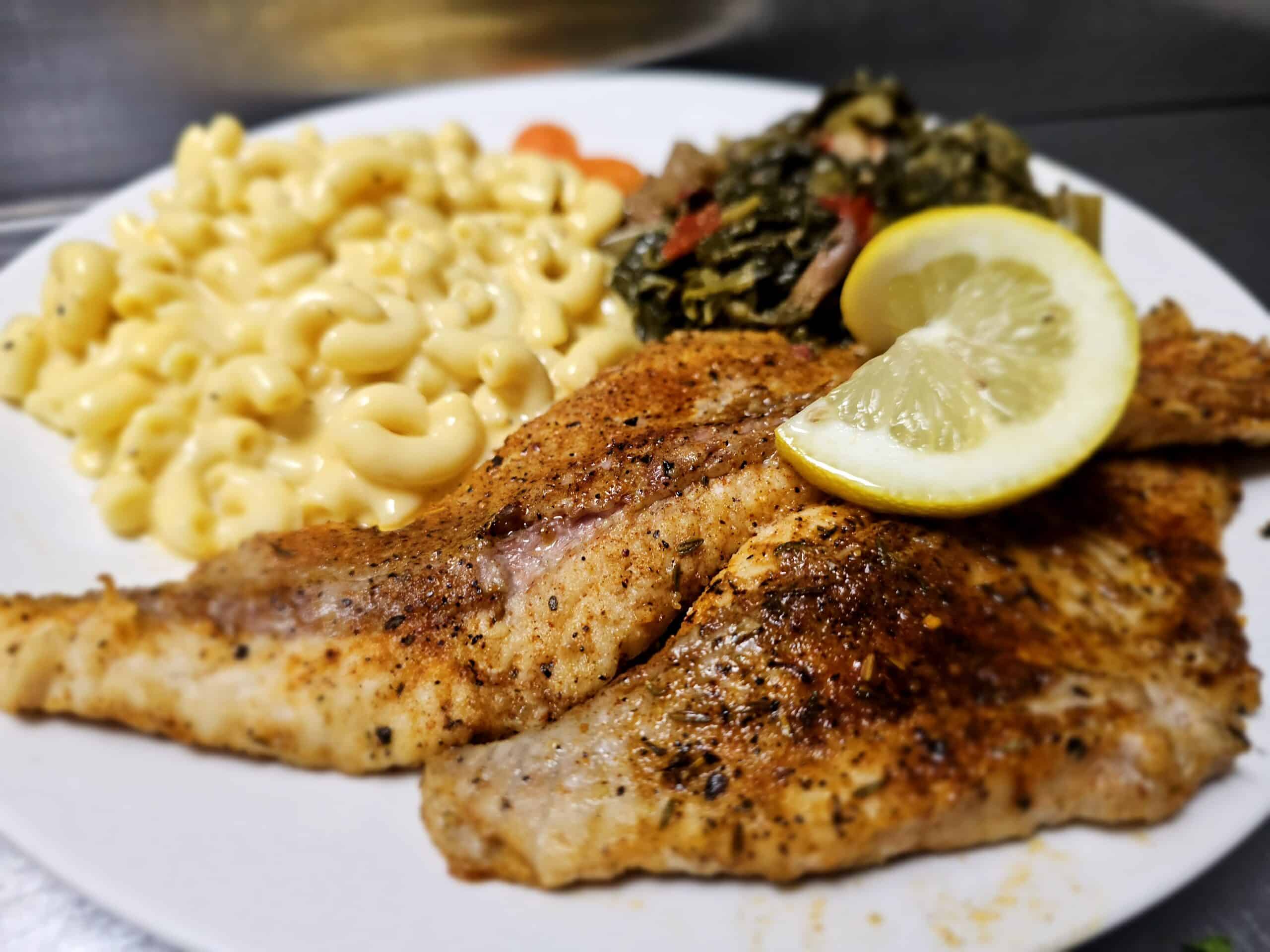 Grilled seasoned chicken with lemon wedge, creamy macaroni and cheese, and greens on a white plate.