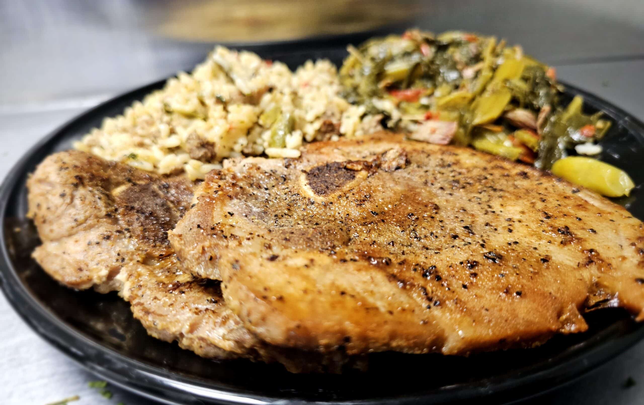 Seasoned grilled pork chop on a plate with rice and sautéed greens on the side, steam rising in the background.