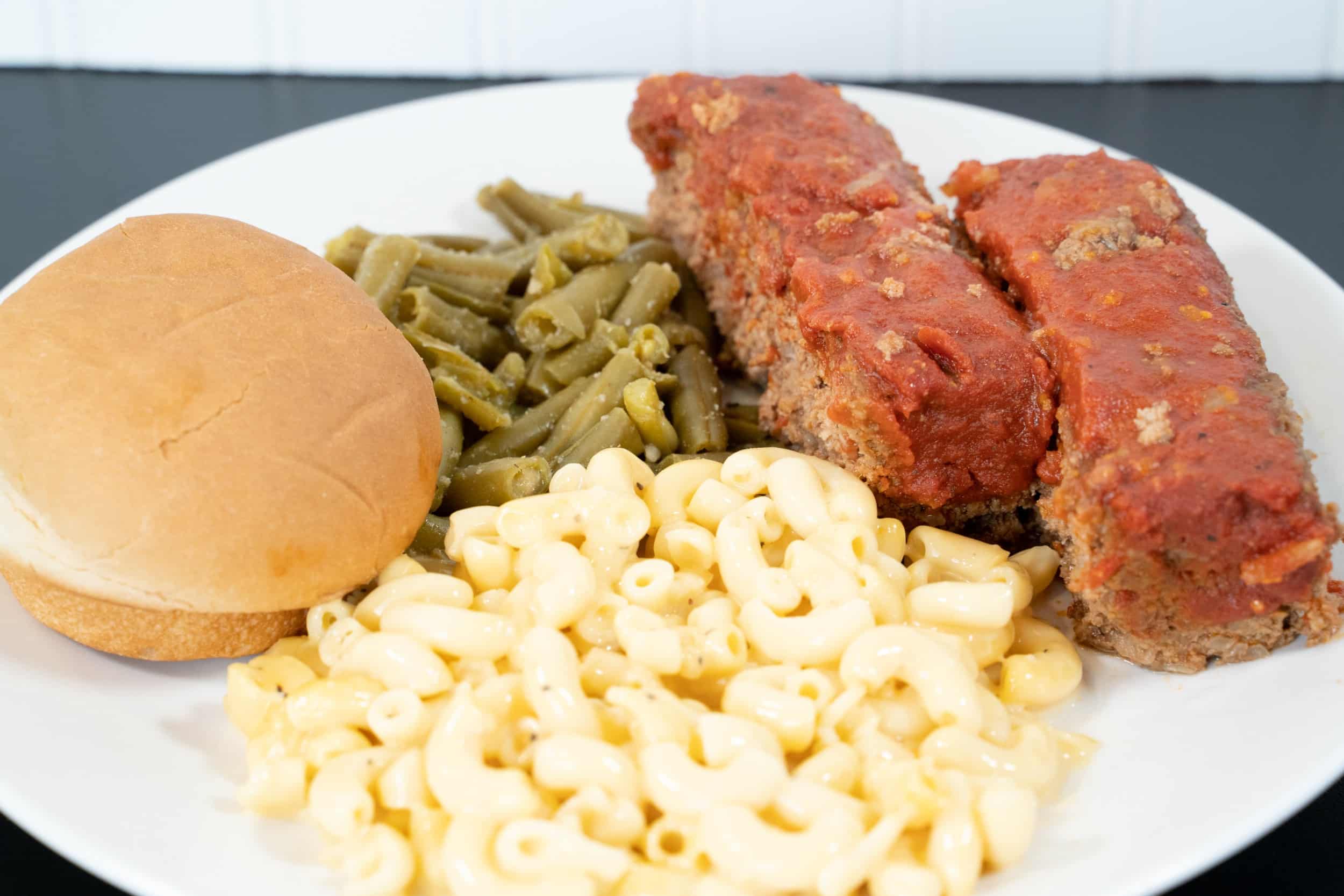 Plate with creamy macaroni and cheese, green beans, meatloaf slices in tomato sauce, and a hamburger bun.