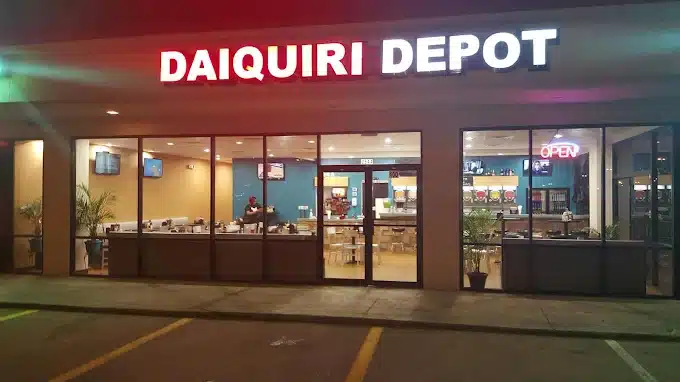 Daiquiri Depot storefront at night with a bright sign, large glass windows showing customers inside and an illuminated OPEN sign.